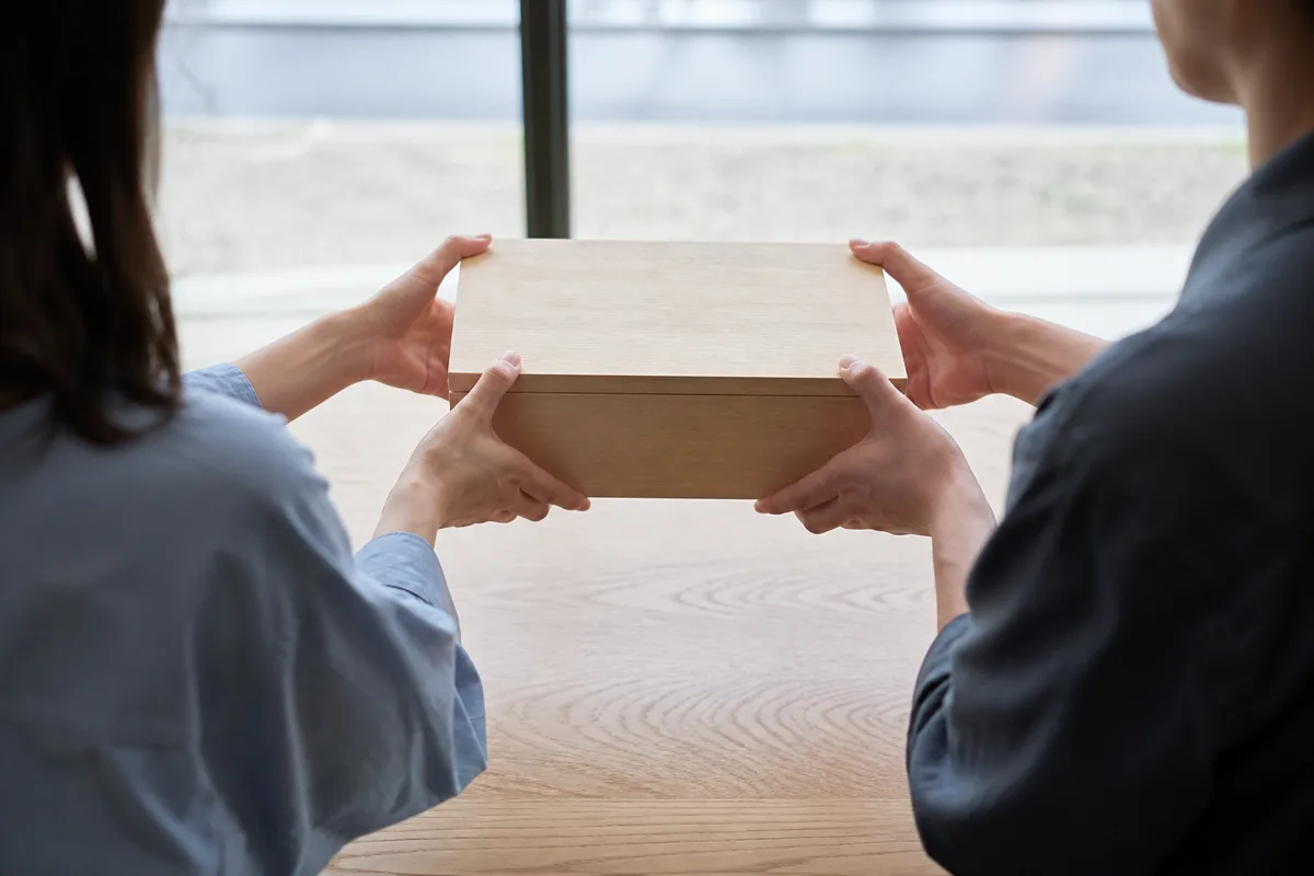 Mains de deux adultes vues de dos passant une boîte en bois clair au-dessus d’une table nue